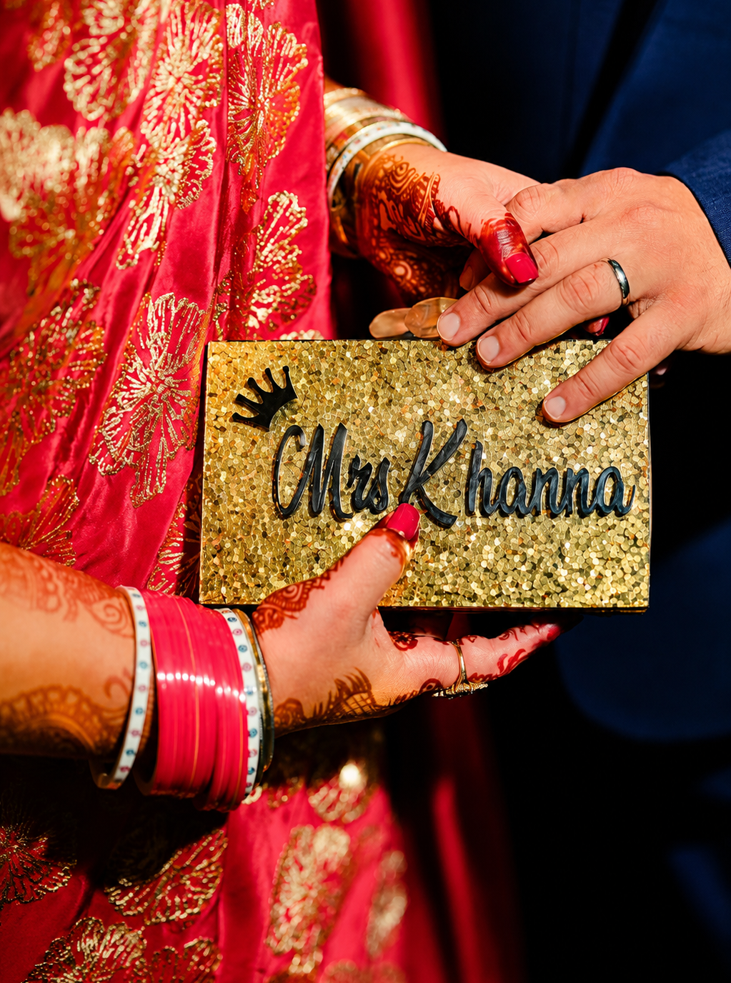 Person holding a gold clutch with 'Mrs Khanna' text, wearing red and gold traditional attire.