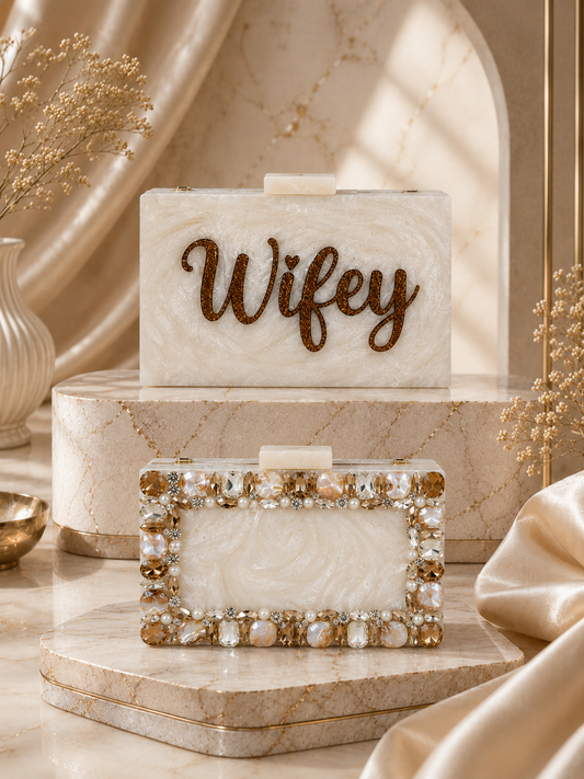 A styled product display of two personalized wedding clutch bags on a marble pedestal. The top bag is rectangular with a cream marbled body and 'Wifey' written in brown script on the front.